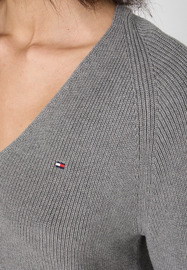 Nahaufnahme einer Person, die einen grauen gerippten V-Ausschnitt-Pullover mit einem kleinen rechteckigen gestickten Logo in Rot, Weiß und Marineblau auf der Brust trägt.
