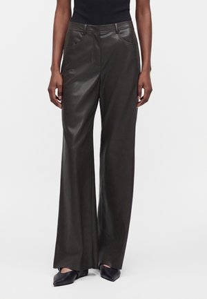 Femme portant un pantalon en cuir noir à jambes larges avec poches avant et des chaussures noires à bout pointu, debout devant un fond uni.