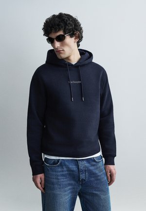 Άνδρας που φοράει μαύρη φούτερ με κορδόνια, μπλε τζιν και μαύρα γυαλιά ηλίου, στέκεται μπροστά σε απλό ανοιχτό γκρι φόντο.