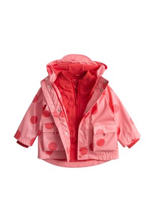 Dziecko w pomarańczowym płaszczu przeciwdeszczowym w grochy, pomarańczowej kurtce, niebieskich dżinsach i brązowych butach balansujące na jednej nodze na jednolitym tle.