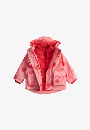 Kind in een oranje regenjas met stippen, oranje jas, blauwe spijkerbroek en bruine laarzen, balancerend op één been tegen een effen achtergrond.
