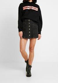 Persona que lleva una sudadera negra de Tommy Jeans, una falda de mezclilla negra con botones en la parte delantera y botas negras hasta el tobillo con hebillas y cremalleras.