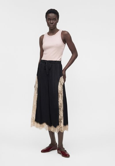 Γυναίκα που φοράει ανοιχτό ροζ αμάνικη μπλούζα, μαύρο παντελόνι με φαρδιά μπατζάκια και μπεζ δαντελωτές λεπτομέρειες, και σκούρα κόκκινα φλατ παπούτσια, στέκεται με το ένα χέρι στην τσέπη.