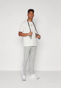 Bela športna majica z rožnato zlatim logotipom, sivo ožane športne hlače z belimi stranskimi črtami in beli športni čevlji. Drži kolebnico.