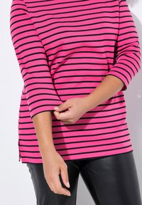 Maglietta a maniche lunghe a righe rosa e nere con fessure laterali, caratterizzata da una texture morbida e un fit rilassato. Indossata con pantaloni neri lucidi.