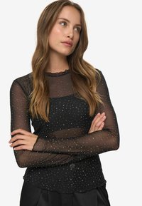 Mlada ženska z dolgimi lasmi, oblečena v črn prosojen zgornji del s srebrnimi bleščicami, z rokami prekrižanimi, ki nekoliko gleda navzgor v desno.