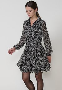 Rochie neagră florală cu mâneci lungi, din material transparent, cu guler butonat și fustă plisată. Are un model cu flori albe și gri.