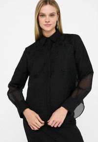 Camicia nera con ricami floreali e motivi paisley, dotata di colletto classico, chiusura frontale con bottoni e maniche lunghe trasparenti.