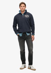 Marineblauer Fleece-Pullover mit Viertelreißverschluss und "SUPERDRY"-Logo, über einem hellblauen Denimhemd getragen, kombiniert mit dunklen Jeans und weißen Sneakers.