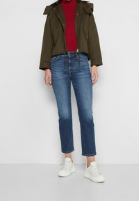 Chaqueta recortada color verde oliva con capucha, suéter de cuello alto rojo, jeans de pierna recta azules y zapatillas blancas. Tejido suave y corte relajado.