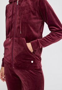Sweat à capuche en velours bordeaux avec une poche avant et des accents en métal argenté. Le tissu a une texture douce et lisse.