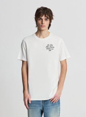 Giovane uomo con capelli castani spettinati, indossa una maglietta bianca con scritta nera sul lato sinistro del petto e jeans blu, in piedi contro uno sfondo grigio.