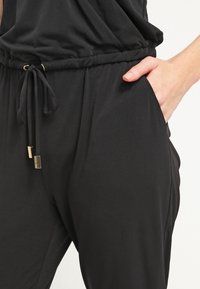 Main gauche dans la poche d'un pantalon noir à taille élastique avec cordon de serrage et extrémités dorées, montrant la texture du tissu et les détails de l'ajustement.