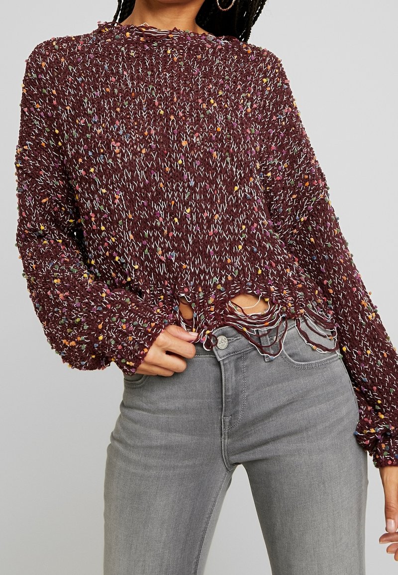 Femme portant un pull en maille multicolore bordeaux avec un ourlet usé et un jean taille haute gris clair, posant avec une main près de la taille.