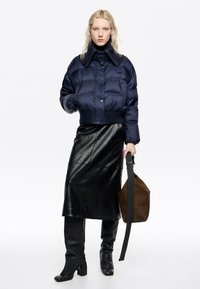 Granatowa kurtka puchowa z wysokim kołnierzem, przednimi kieszeniami i prążkowanym dołem. Połączona z błyszczącą czarną skórzaną spódnicą i brązową torebką na ramię.