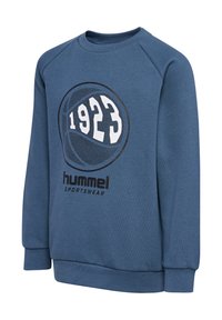 Niebieski sweter z okrągłym kołnierzem, z raglanowymi rękawami, z okrągłą grafiką z napisem "1923" oraz "hummel sportswear" w czarnym druku. Materiał mieszanki bawełnianej.