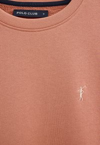 Zbliżenie na różową, zakurzoną bluzę z okrągłym dekoltem, z czarną metką "Polo Club" i małym białym haftem golfisty z kijem.