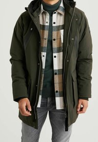 Ciemnozielona ocieplana kurtka na zamek błyskawiczny z kapturem. Na niej warstwowa koszula w kratę w odcieniach beżu, szarości i zieleni, pod którą widoczny jest ciemnozielony sweter.