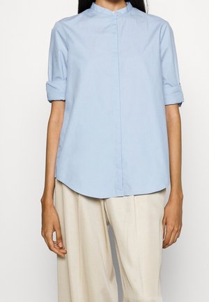 Femme portant un chemisier bleu clair à manches courtes avec col mao et un pantalon beige taille haute à jambes larges sur fond blanc.