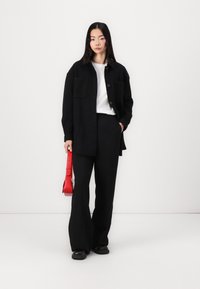 Zwarte oversized button-up shirt met twee zakken, gecombineerd met zwarte wijde broek en een rode schoudertas. Gladde stof, minimalistisch ontwerp.