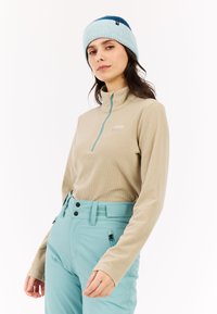 Licht beige langmouwtop met een kwart-rits en teal accenten, gecombineerd met lichtblauwe high-waisted broeken en een meerkleurige gebreide muts.