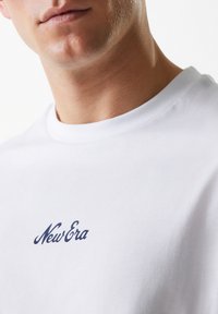 Bijela pamučna majica s okruglim izrezom na kojoj je na prsima izvezen tamnoplavi "New Era" logo. Glatka tekstura i prilagođeni dizajn.