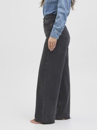 Kvinne som har på seg høytlivede svarte viddejeans og en langermet blå denimskjorte, som står barbent foran en ensfarget hvit bakgrunn.