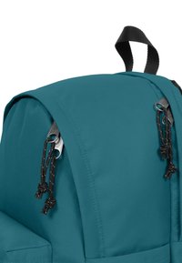 Turkos ryggsäck med en slät yta, rund form, svarta detaljer och snörstängning upptill. Remmarna är förstärkta.