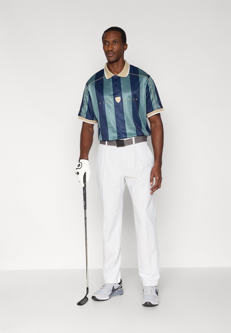 Completo da golf composto da una polo leggera a righe turchesi e blu navy con colletto crema, abbinata a pantaloni bianchi e scarpe da ginnastica grigie.