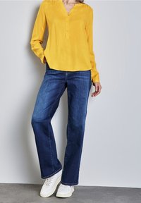 Persona che indossa una blusa gialla a maniche lunghe, jeans blu e sneaker bianche, in piedi contro un muro grigio chiaro con una mano in tasca.