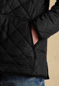 Czarna kurtka pikowana z diamentowym przeszyciem, z ręką w przedniej kieszeni, w połączeniu z niebieskimi dżinsami.