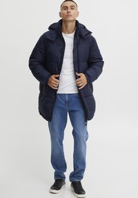 Marineblå pufferjakke med hette, quiltet design, frontlukking med glidelås og elastiske mansjetter. Kombinert med blå jeans og svarte sneakers.