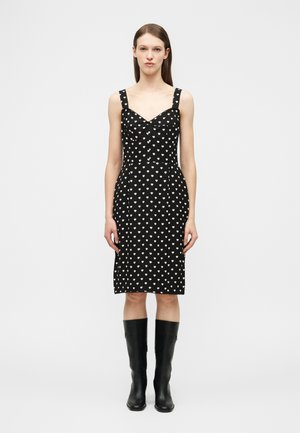 Mujer de pie con un vestido negro hasta la rodilla con patrones de corazones blancos y botas negras hasta la rodilla, sobre un fondo gris liso.