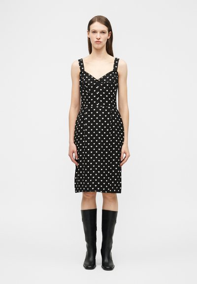 Mujer de pie con un vestido negro hasta la rodilla con patrones de corazones blancos y botas negras hasta la rodilla, sobre un fondo gris liso.