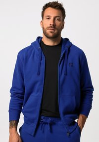 Sudadera con cremallera azul en una tela suave, con un logo negro en el pecho, que cuenta con una capucha forrada y bolsillos frontales, combinada con una camiseta negra debajo.