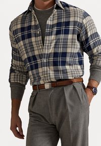 Mies pukeutuneena siniseen ja beigeen ruudulliseen napilliseen paitaan harmaan neuleen päällä, harmaat laskospöksyt, ruskea nahkavyö ja ruskealla remmillä varustettu kello.