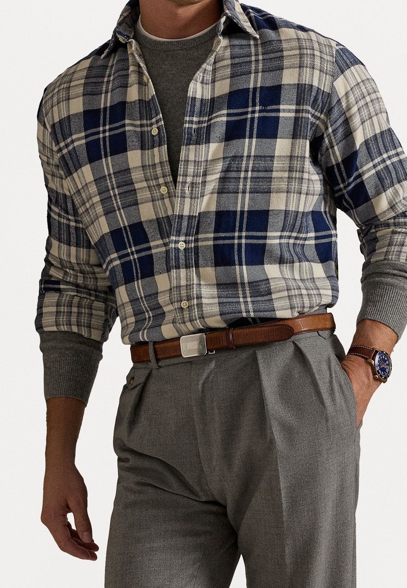 Mies pukeutuneena siniseen ja beigeen ruudulliseen napilliseen paitaan harmaan neuleen päällä, harmaat laskospöksyt, ruskea nahkavyö ja ruskealla remmillä varustettu kello.