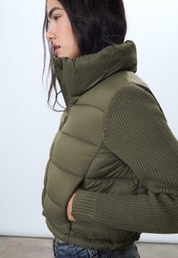 Padded vest v olivovej farbe s vysokým golierom, ktorý má textúrované pletené rukávy a bočné vrecká, navrhnutý pre teplo a pohodlie.