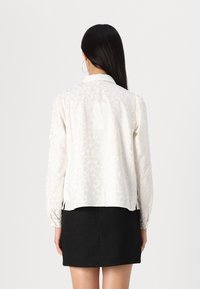 Blouse blanche avec un motif texturé en forme de cœur, manches longues et col, associée à une jupe noire texturée.