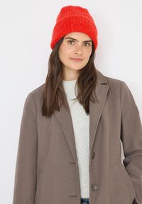 Cappotto di lana marrone con rever a punta, due bottoni e tasche laterali. Maglione grigio chiaro texturizzato sotto e berretto di lana rosso brillante.