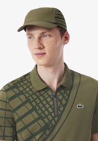 Olijfgroene polo met een halve rits, voorzien van een geometrisch patroon op de bovenkant. Een bijpassende olijfkleurige pet maakt de outfit compleet.