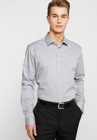 Camicia grigia a maniche lunghe con colletto classico, texture sottile e bottoni bianchi, abbinata a pantaloni neri e una cintura.