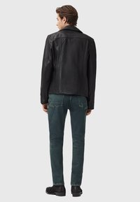 Černá kožená bunda s hladkým povrchem, s klasickým límcem a precizním šitím, ve spojení s tmavozelenými džíny a černými botami.