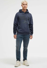 Navy hoodie with graphic print, drawstring hood, and zippered side pockets, paired with blue jeans and light sneakers. Smooth texture.