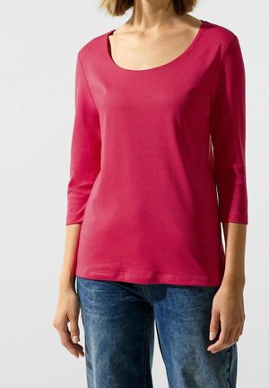 Kobieta ubrana w gładki różowy top z rękawami do połowy łokcia i niebieskie dżinsy, stojąca na jasnym tle.