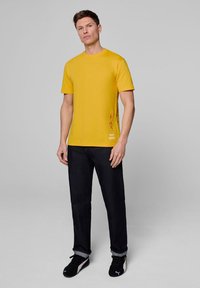 Żółta, krótko rękawowa koszulka z bawełny, z czerwonymi akcentami graficznymi i logo na dolnym brzegu, zestawiona z ciemnymi dżinsami i czarnymi sneakersami.