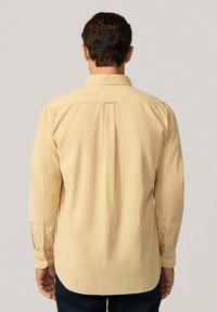 Camisa de manga larga en amarillo claro, hecha de una tela suave. Presenta un cuello con botones y un pliegue en la espalda, con puños abotonados y dobladillo redondeado.