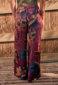 Pantalon à jambes larges en tissu bordeaux profond, avec des motifs floraux colorés en or, bleu et rouge. Surface texturée avec une taille élastique.