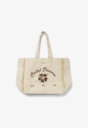 Sac fourre-tout en toile beige avec doubles poignées, bords verticaux effilochés, et texte marron « Costal Dreams feel the sea » entourant un motif de fleur d’hibiscus.