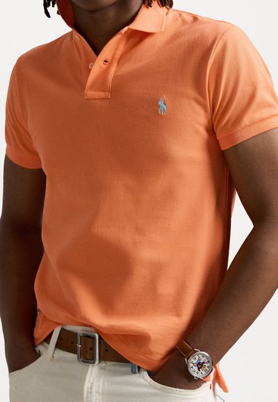 Oranžs polo krekls, kas izgatavots no teksturētas kokvilnas, ar divu pogu aizdari un mazu zilu izšuvumu logotipu. Nēsāts ar bēša jostu un pulksteni.
